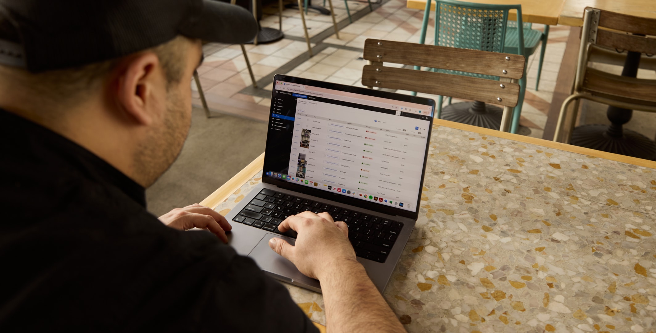 Person sitting at a table, using a laptop in a cafe. The screen displays a list with columns, possibly a spreadsheet or data table.